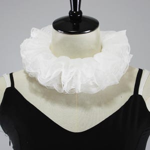 May include: A white ruffled lace neck collar featuring a layered, pleated design with delicate scalloped edges. The accessory is displayed around the neck of a mannequin, showcasing a voluminous, circular silhouette that sits above a black garment.