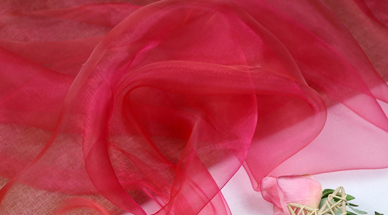 May include: A close-up of a bright red sheer fabric, with a soft, flowing texture. The fabric is draped and folded, creating a delicate and airy appearance.