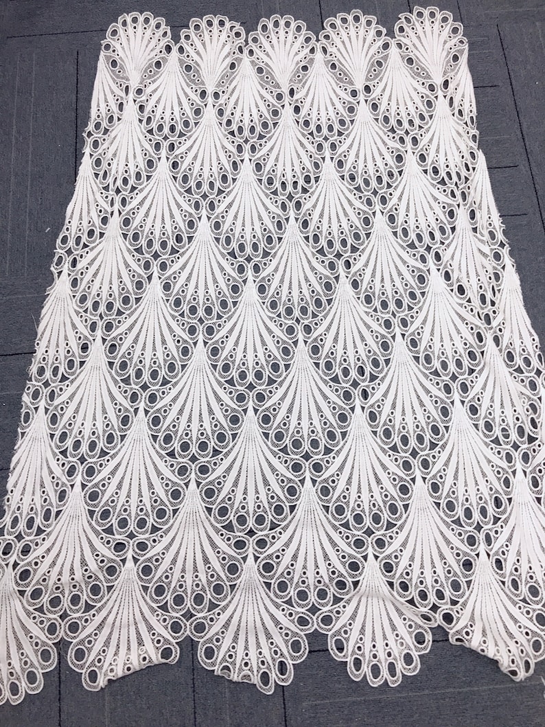 May include: White lace fabric with a repeating pattern of stylized peacock feathers. The delicate design features openwork circles and curved lines, creating a textured, airy appearance. The fabric is likely used for apparel or decorative purposes.