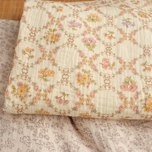 May include: Cream-colored quilted fabric with a floral pattern. The design features a repeating pattern of oval frames with colorful flowers in shades of pink, yellow, and purple. The fabric is folded, showing the texture.