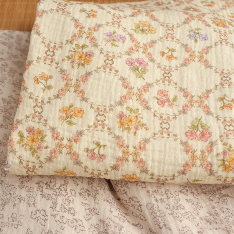 May include: Cream-colored quilted fabric with a floral pattern. The design features a repeating pattern of oval frames with colorful flowers in shades of pink, yellow, and purple. The fabric is folded, showing the texture.