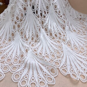 May include: White lace fabric with a peacock feather design. The fabric features a repeating pattern of oval shapes and curved lines, creating a delicate and intricate texture. The lace is likely used for apparel or home decor.