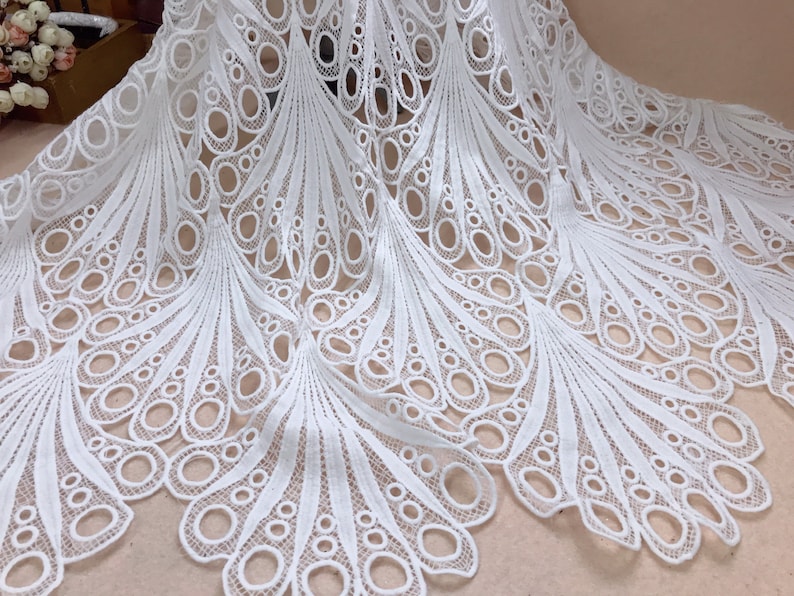 May include: White lace fabric with a peacock feather design. The fabric features a repeating pattern of oval shapes and curved lines, creating a delicate and intricate texture. The lace is likely used for apparel or home decor.