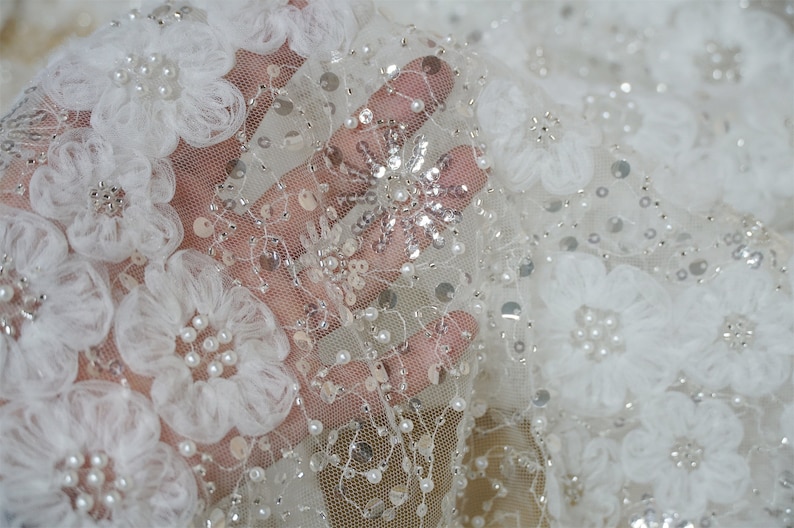 以下が含まれることがあります： 白いチュール生地に、3Dの花のアップリケとパールとスパンコールの装飾が施されています。