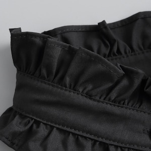 May include: Close-up of a black garment featuring a ruffled collar and a button closure. The fabric appears to be a smooth, possibly satin-like material. The stitching is visible, adding detail to the design.