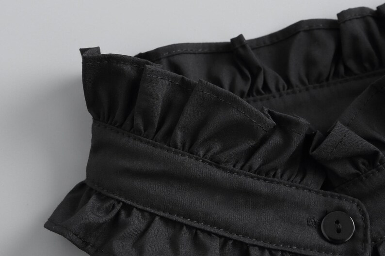May include: Close-up of a black garment featuring a ruffled collar and a button closure. The fabric appears to be a smooth, possibly satin-like material. The stitching is visible, adding detail to the design.