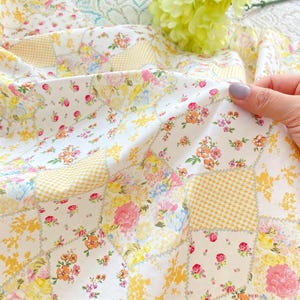 Puede incluir: Un edredón de patchwork con un estampado floral en tonos rosa, amarillo y naranja sobre un fondo blanco. El edredón tiene una mezcla de cuadrados florales y de cuadros vichy. La tela es suave y parece ser de algodón.