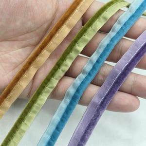Op de afbeelding: Vier stroken fluwelen biesband in de kleuren goudgeel, olijfgroen, lichtblauw en lavendel. Elke strook heeft aan één kant een zachte, pluizige fluwelen textuur en een platte, geweven stoffen rand met zichtbare stiksels langs de naad.