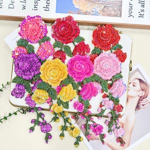 Puede incluir: Apliques florales bordados en tonos rosa, rojo, amarillo y morado con hojas verdes. Los apliques están adornados con lentejuelas y abalorios, y están dispuestos sobre una superficie blanca.