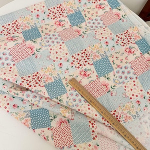 May include: A patchwork fabric with a floral pattern in pink, red, and yellow, interspersed with light blue squares with white polka dots. A wooden ruler is placed on the fabric. The fabric is likely cotton.