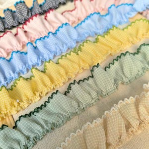 May include: A collection of ruffled fabric trim strips featuring a small gingham check pattern in various colors including black, pink, blue, yellow, green, and beige. Each strip has a decorative scalloped edge stitched in a contrasting thread.