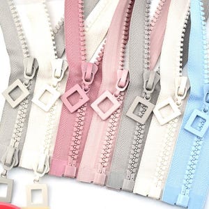 May include: A collection of colorful zippers in various shades, including gray, white, pink, and blue. Each zipper features a square-shaped pull tab. The zippers are arranged side by side, showcasing their different colors and designs.