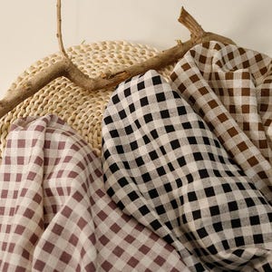 May include: Close-up of fabric swatches in various colours and patterns. The fabrics feature a classic gingham check design in shades of black and white, brown and white, and mauve and white. The fabrics are draped over a woven mat.