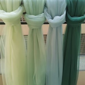 May include: Four lengths of sheer fabric in varying shades of green, from pale lime to deep forest, are draped over a metal bar. The fabric is gathered and tied, creating a layered effect. The soft, flowing material suggests a decorative or craft application.