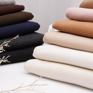 Peut inclure: Plusieurs piles de tissus pliés de différentes couleurs : gris, marron, marine et noir à gauche, beige, rose et beige clair à droite. Le tissu semble doux, peut-être de la laine ou un matériau similaire. L'image est bien éclairée.