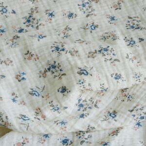 May include: A white fabric with a small floral pattern in shades of blue and pink. The fabric is soft and textured, with a slightly crinkled appearance.