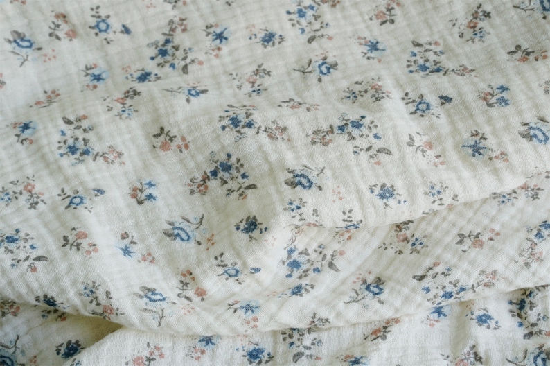 May include: A white fabric with a small floral pattern in shades of blue and pink. The fabric is soft and textured, with a slightly crinkled appearance.