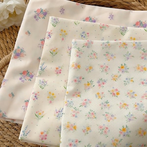 Pode incluir: Três tecidos dobrados com estampas florais. O tecido superior é de cor creme claro com grandes buquês florais em rosa, roxo e azul. O tecido do meio é branco com pequenos buquês florais em rosa, amarelo e azul. O tecido inferior é branco com pequenos buquês florais.