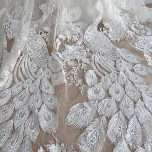 Puede incluir: Tela de encaje blanca con cuentas con un diseño de pavo real. La tela es transparente y tiene un aspecto delicado y femenino.