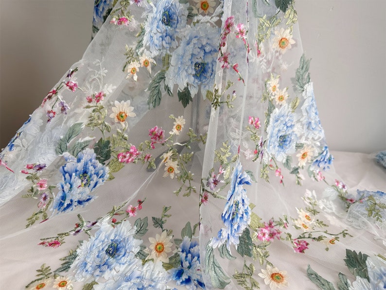 Puede incluir: Tela blanca transparente con un patr&oacute;n floral bordado. Las flores son azules, rosas y amarillas. La tela es delicada y tiene una textura suave.