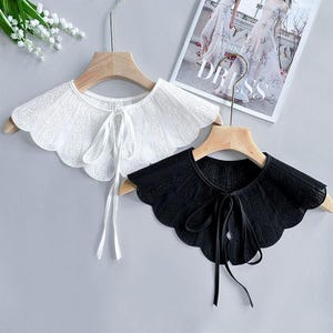 Könnte beinhalten: Zwei Spitzenkragen-Accessoires, einer weiß und einer schwarz, auf Holzbügeln präsentiert. Beide Kragen haben einen gewellten Rand und einen Krawattenverschluss. Der weiße Kragen hat ein zartes Blumenmuster, während der schwarze Kragen ein ähnliches, aber anderes Spitzendesign hat.