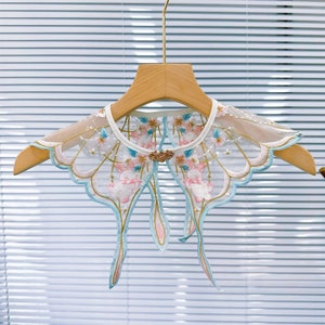 May include: A delicate white lace collar with blue and pink floral embroidery and a gold button detail. The collar is hanging on a wooden hanger.