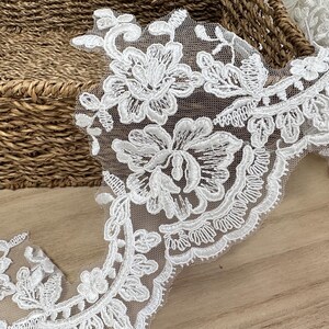 May include: Detailed shot of white floral lace trim featuring embroidered flowers and leaves. The lace is displayed on a light brown wooden surface, with a woven basket in the background, highlighting the delicate craftsmanship.