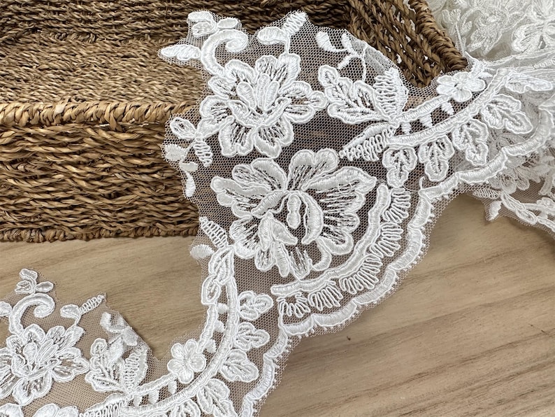 May include: Detailed shot of white floral lace trim featuring embroidered flowers and leaves. The lace is displayed on a light brown wooden surface, with a woven basket in the background, highlighting the delicate craftsmanship.
