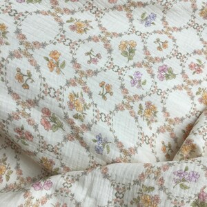 May include: Cream-colored fabric with a repeating floral pattern. Each floral arrangement is encircled by a delicate garland of leaves and flowers in shades of pink, yellow, orange, and purple. The fabric has a subtle, textured appearance.