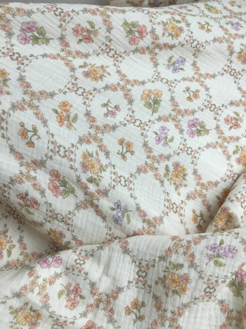 May include: Cream-colored fabric with a repeating floral pattern. Each floral arrangement is encircled by a delicate garland of leaves and flowers in shades of pink, yellow, orange, and purple. The fabric has a subtle, textured appearance.