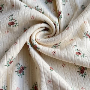 May include: Close-up of a cream-colored fabric with a ribbed texture and a floral pattern. The flowers are a mix of red and green, scattered across the material. The fabric is folded, creating a swirling effect.