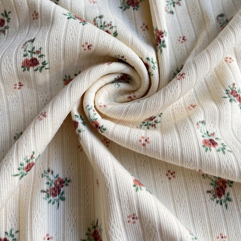May include: Close-up of a cream-colored fabric with a ribbed texture and a floral pattern. The flowers are a mix of red and green, scattered across the material. The fabric is folded, creating a swirling effect.