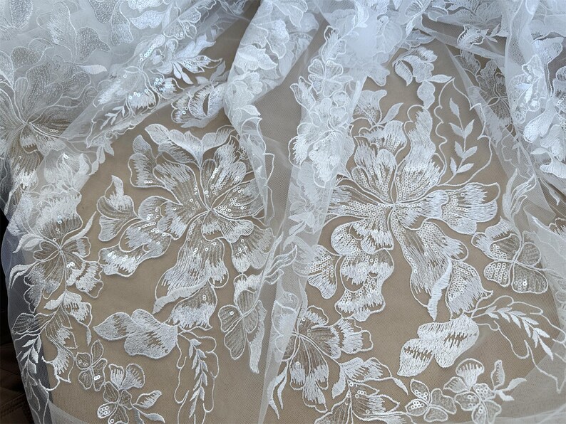 May include: White floral lace fabric with a delicate pattern of flowers and leaves. The lace is embellished with sequins, adding a touch of sparkle.