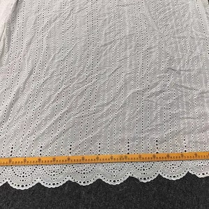May include: White cotton fabric with a delicate eyelet pattern. The fabric has a scalloped edge and is measured with a ruler in inches.