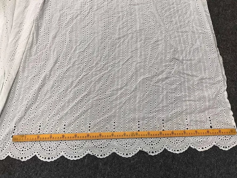 May include: White cotton fabric with a delicate eyelet pattern. The fabric has a scalloped edge and is measured with a ruler in inches.