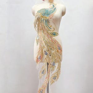 May include: A large peacock-shaped applique featuring intricate embroidery in shades of turquoise, gold, and soft pink. The design is embellished with shimmering sequins and beads, forming a detailed bird body and long, flowing tail feathers.