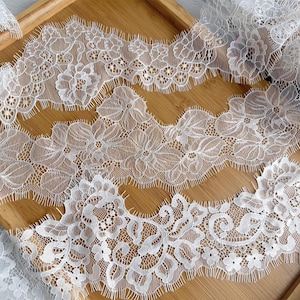 May include: A length of white lace trim featuring a detailed floral pattern with scalloped edges and delicate eyelash fringe. The intricate mesh design displays repeating flower motifs connected by fine threads, creating a continuous decorative border.