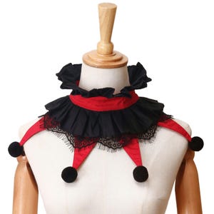 May include: A red and black neck ruff featuring a pleated black fabric collar, a red fabric base, and black lace trim. The accessory includes three pointed fabric extensions, each finished with a small, round, black pom-pom at the tip.