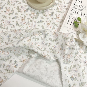 Op de afbeelding: Een witte stof met een herhalend patroon van kleine ganzen met blauwe mutsjes, afgewisseld met groene takjes en kleine blauwe bloemetjes. Het textiel heeft een zachte, geplooide textuur die de gedetailleerde illustratie van de ganzen toont.