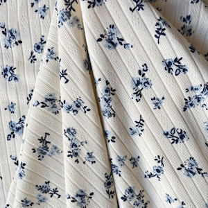 May include: Close-up of a cream-colored fabric with a ribbed texture. The fabric is adorned with a pattern of small, blue floral clusters. The flowers have delicate petals and are scattered across the material, creating a soft, feminine aesthetic.
