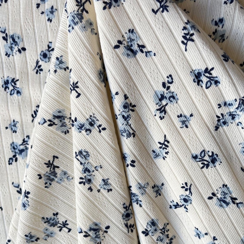 May include: Close-up of a cream-colored fabric with a ribbed texture. The fabric is adorned with a pattern of small, blue floral clusters. The flowers have delicate petals and are scattered across the material, creating a soft, feminine aesthetic.