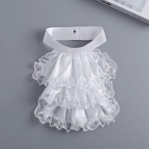 Pode incluir: Um colarinho jabot branco removível com três camadas de tecido acetinado e acabamento em renda delicada nas bordas. A tira do pescoço inclui um fecho de botão de pressão em metal prateado na parte central traseira para fixação segura.