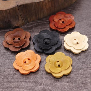 Könnte beinhalten: Eine Sammlung von fünf blumenförmigen Knöpfen aus Holz, die jeweils zwei mittige Löcher aufweisen. Die Knöpfe sind in verschiedenen Farben gehalten, darunter Dunkelbraun, Rotbraun, Schwarz, Hellbeige, Orange und Senfgelb.