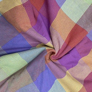 Puede incluir: Primer plano de una colorida tela a cuadros con una textura tejida. El patrón presenta una cuadrícula de líneas que se cruzan en tonos de morado, rojo, amarillo y azul. La tela está doblada y drapeada, creando sombras y profundidad.