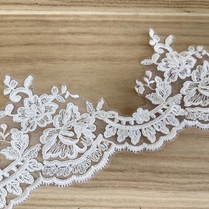 May include: Ivory floral lace trim with a scalloped edge. The delicate lace features detailed floral and leaf patterns, ideal for embellishing apparel or craft projects. The trim is set against a light brown wooden surface.