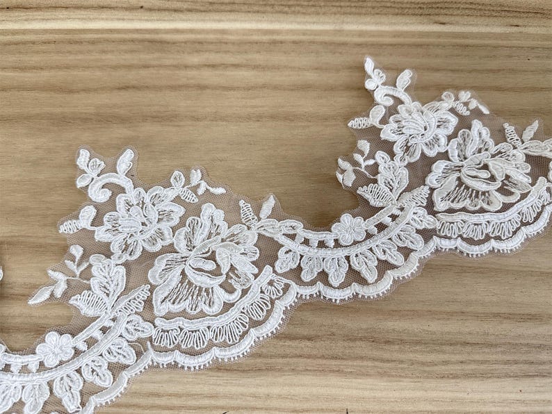 May include: Ivory floral lace trim with a scalloped edge. The delicate lace features detailed floral and leaf patterns, ideal for embellishing apparel or craft projects. The trim is set against a light brown wooden surface.