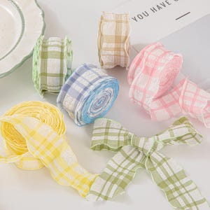 Op de afbeelding: Assortiment van kleurrijke geruite linten in geel, groen, blauw, roze en beige tinten. Sommige linten zijn opgerold, andere zijn in strikken gevormd. De linten zijn gemaakt van een transparante stof met een getextureerd oppervlak.