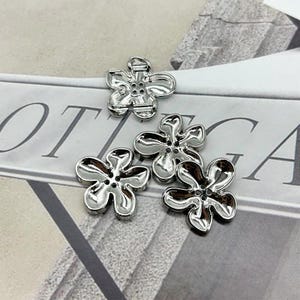 Pode incluir: Um conjunto de botões prateados em forma de flor. Cada botão tem um acabamento brilhante e um design de quatro pétalas com pequenos orifícios para costura. Os botões estão dispostos sobre uma superfície cinza claro.