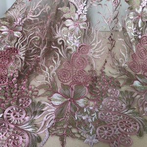 May include: Close-up of a floral lace fabric. The fabric features intricate embroidery in shades of pink, gold, and green. The design includes flowers, leaves, and swirling patterns, creating a delicate and ornate texture. The lace is likely used for apparel or decorative purposes.