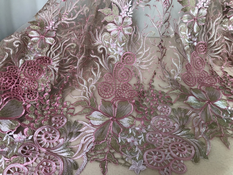 May include: Close-up of a floral lace fabric. The fabric features intricate embroidery in shades of pink, gold, and green. The design includes flowers, leaves, and swirling patterns, creating a delicate and ornate texture. The lace is likely used for apparel or decorative purposes.